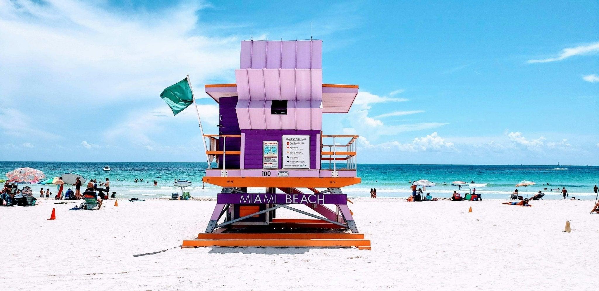 A vibrant, sunny day at Miami Beach featuring a colorful purple and orange lifeguard tower with a green flag, set against turquoise ocean waters and clear blue skies. Beachgoers relax under umbrellas and swim in the ocean, capturing the lively atmosphere