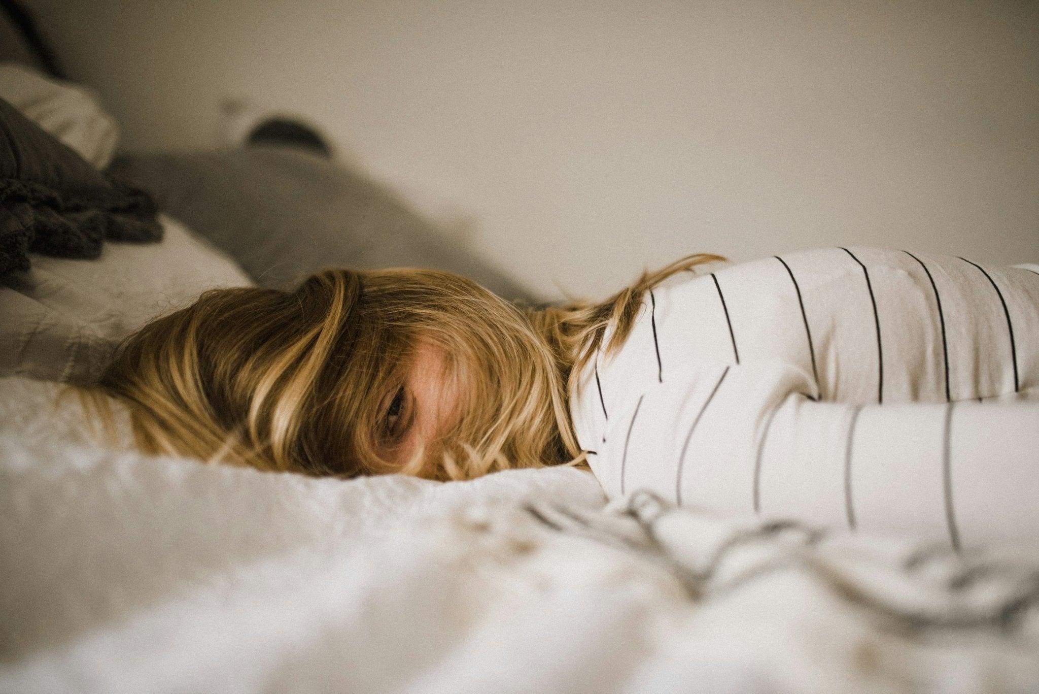 A relaxed person lying on a bed, partially hiding their face behind their tousled blonde hair. They’re wearing a comfortable white shirt with thin black stripes. The overall atmosphere is cozy and casual, suggesting a restful or lazy day indoors.